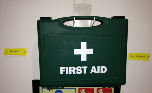 Une image d'un kit de premier secours sur un mur entouré de balises disant ‘Premier secours’ et ‘/Premier secours’